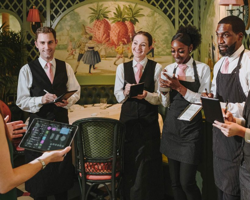 Restaurant employees with notepads are standing in a circle, listening to someone with an iPad speak