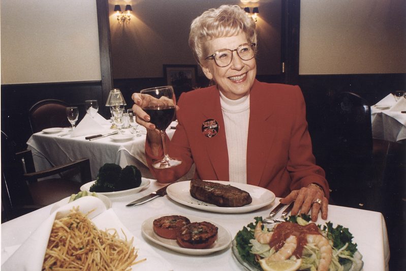 Ruth Fertel at table with red wine, steak, fries, and shrimp salad