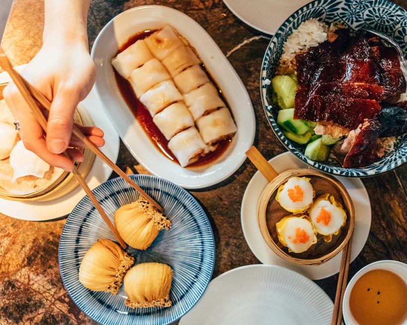A table laden with Chinese dim sum and human hands picking up with chopsticks