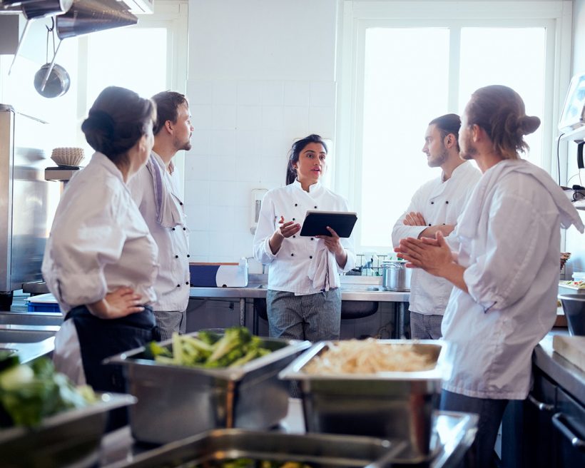 Female chef holding a digital tablet while discussing with team in kitchen