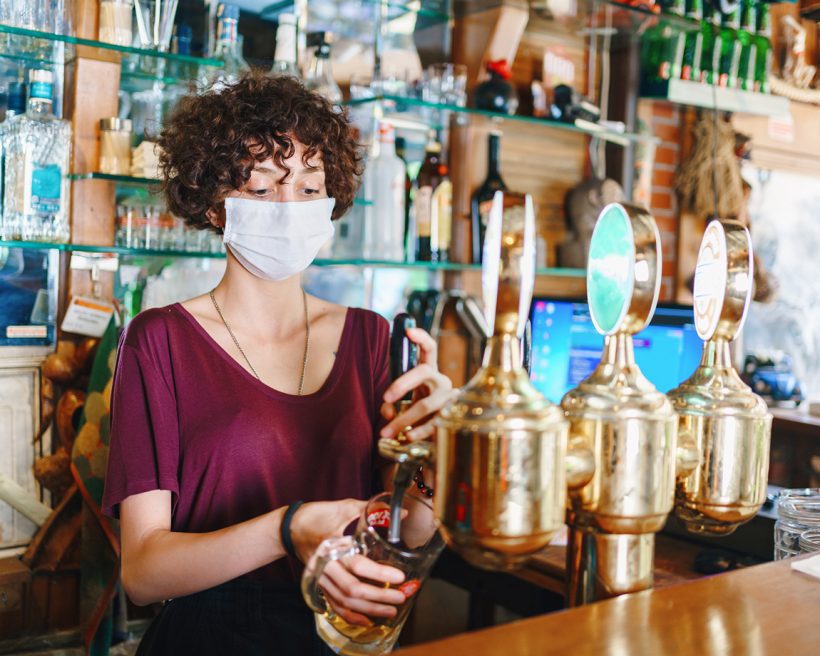 A masked bartender pours a draft beer