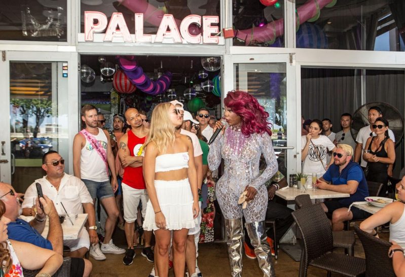 A drag performer talks into a microphone next to a diner.