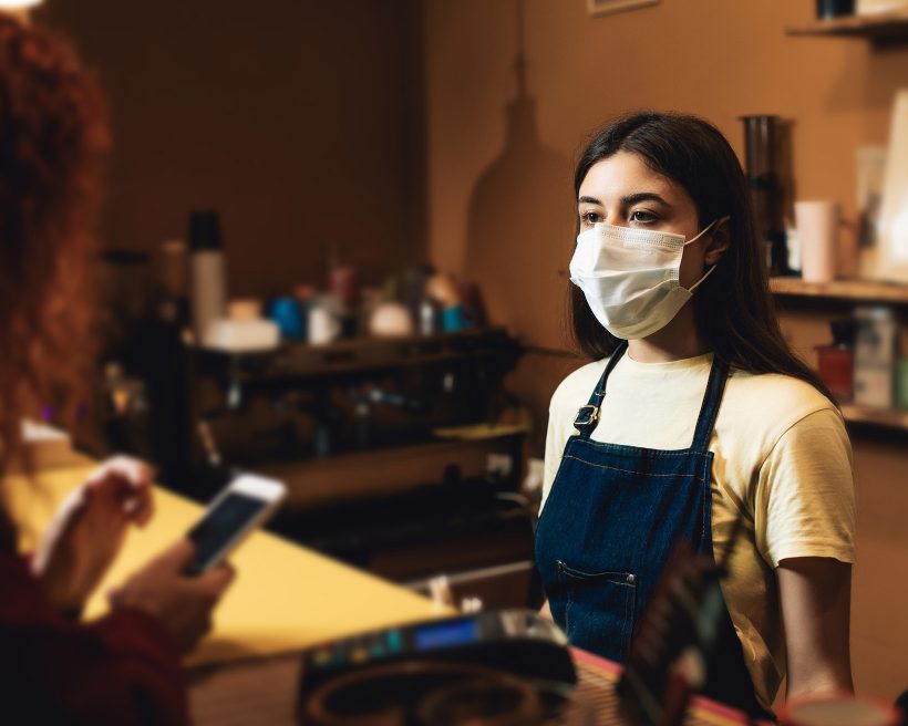 Young woman with facial medical personal protective equipment. Lady is working as barista in cafe. Portrait with facemask, girl is preparing coffee for client