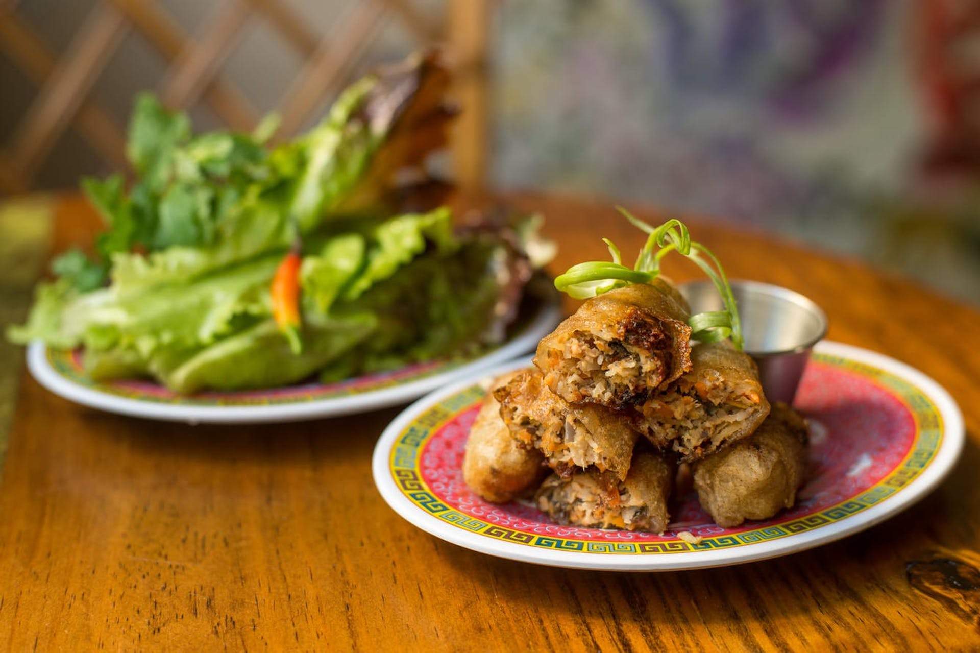 Two plates on a wooden table. One has green salad leaves on one side and the other has crispy spring rolls on it.