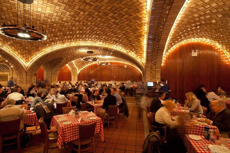 Under the rotund ceilings of the Grand Central Oyster Bar in New York City, diners dig into oysters and cocktails at tables with red-checkered tablecloths.