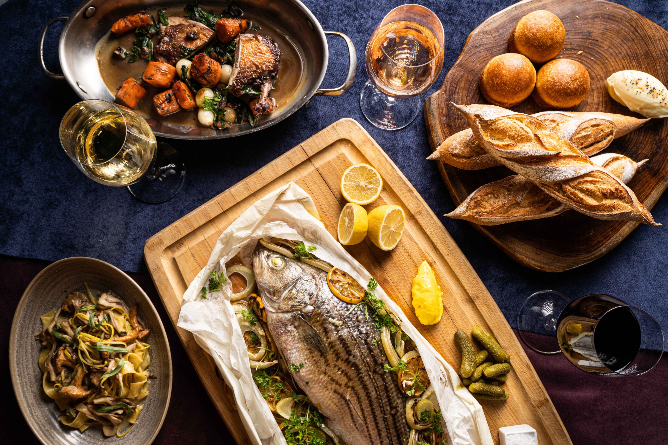 A spread of French classics at Bistro Vendôme in Denver including whole-striped sea bass en papillote, a bread basket, and several glasses of wine