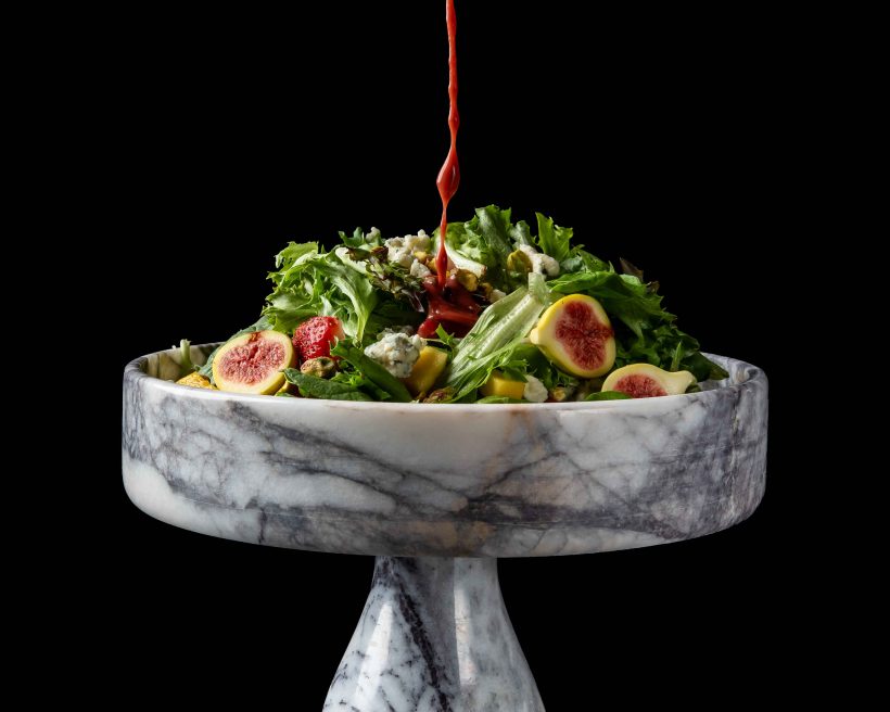 A hand pours raspberry maple balsamic dressing over a salad of mixed greens, strawberries, and figs at GAIA Steakhouse in New Orleans.