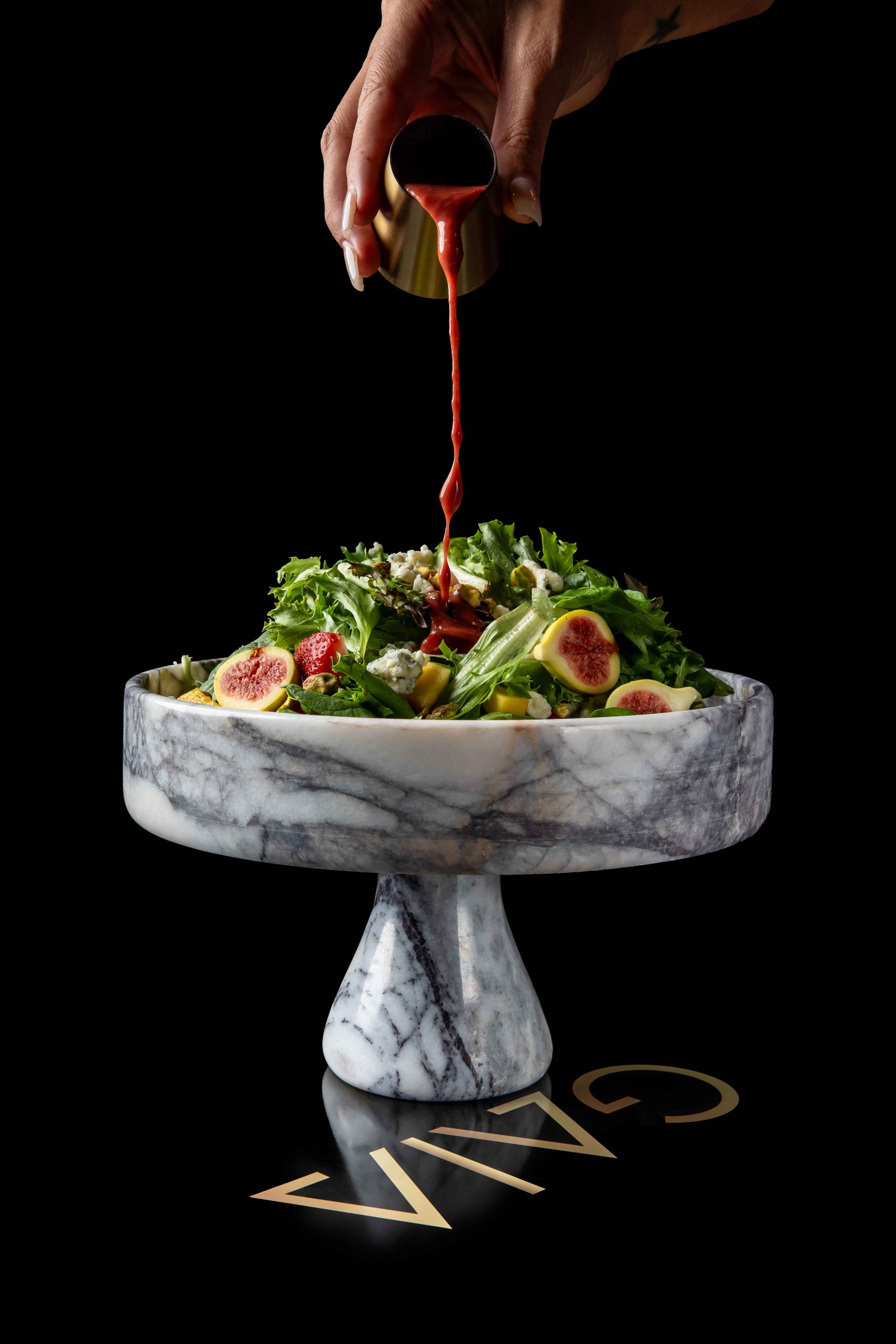A hand pours raspberry maple balsamic dressing over a salad of mixed greens, strawberries, and figs at GAIA Steakhouse in New Orleans.