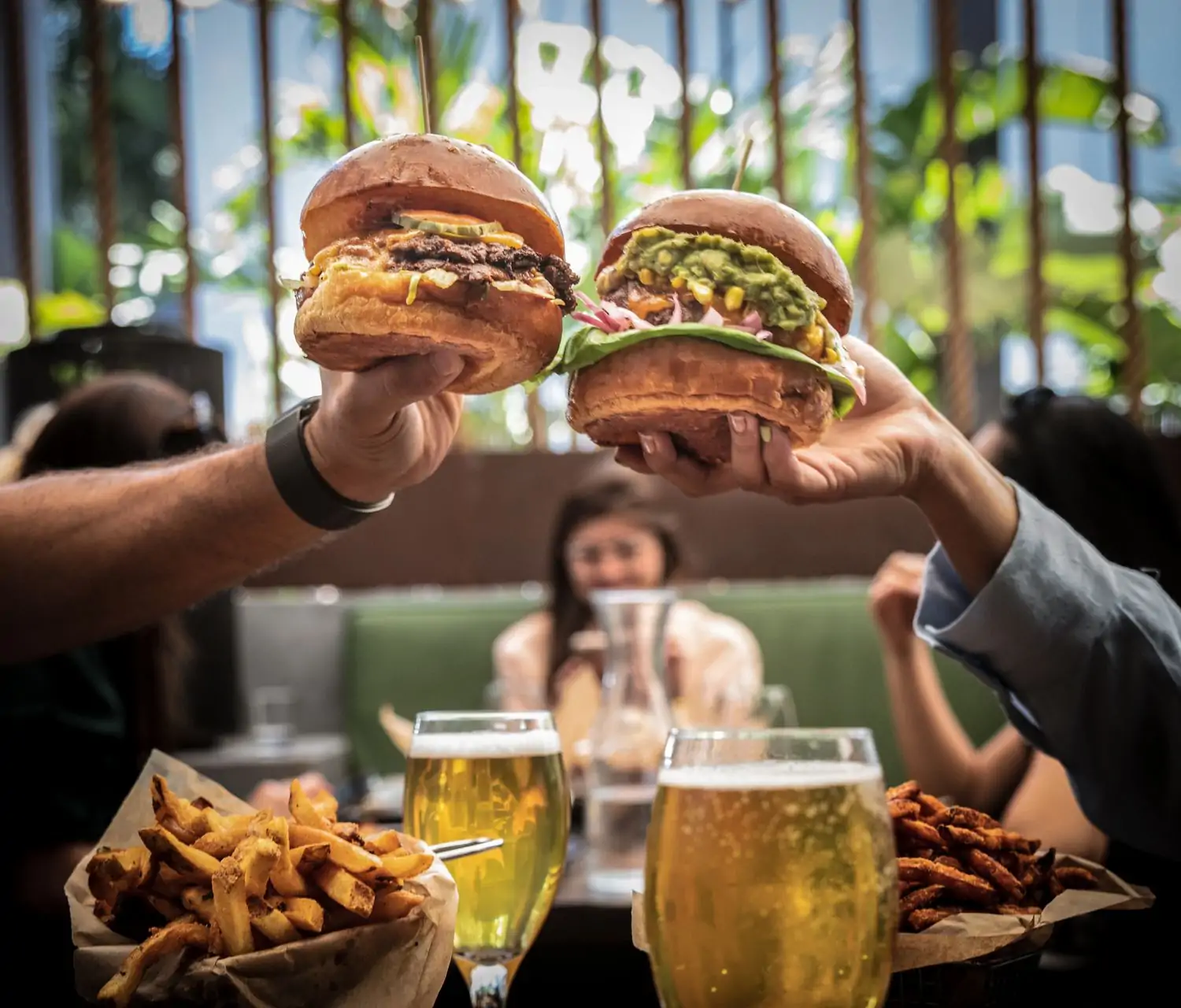 Two people toasting with their burgers from Le Chick, one of the best restaurants in Wynwood, Miami. The hearty burgers are topped with cheese and onions, and one has guacamole added. Also pictured are baskets of fries and two beers.
