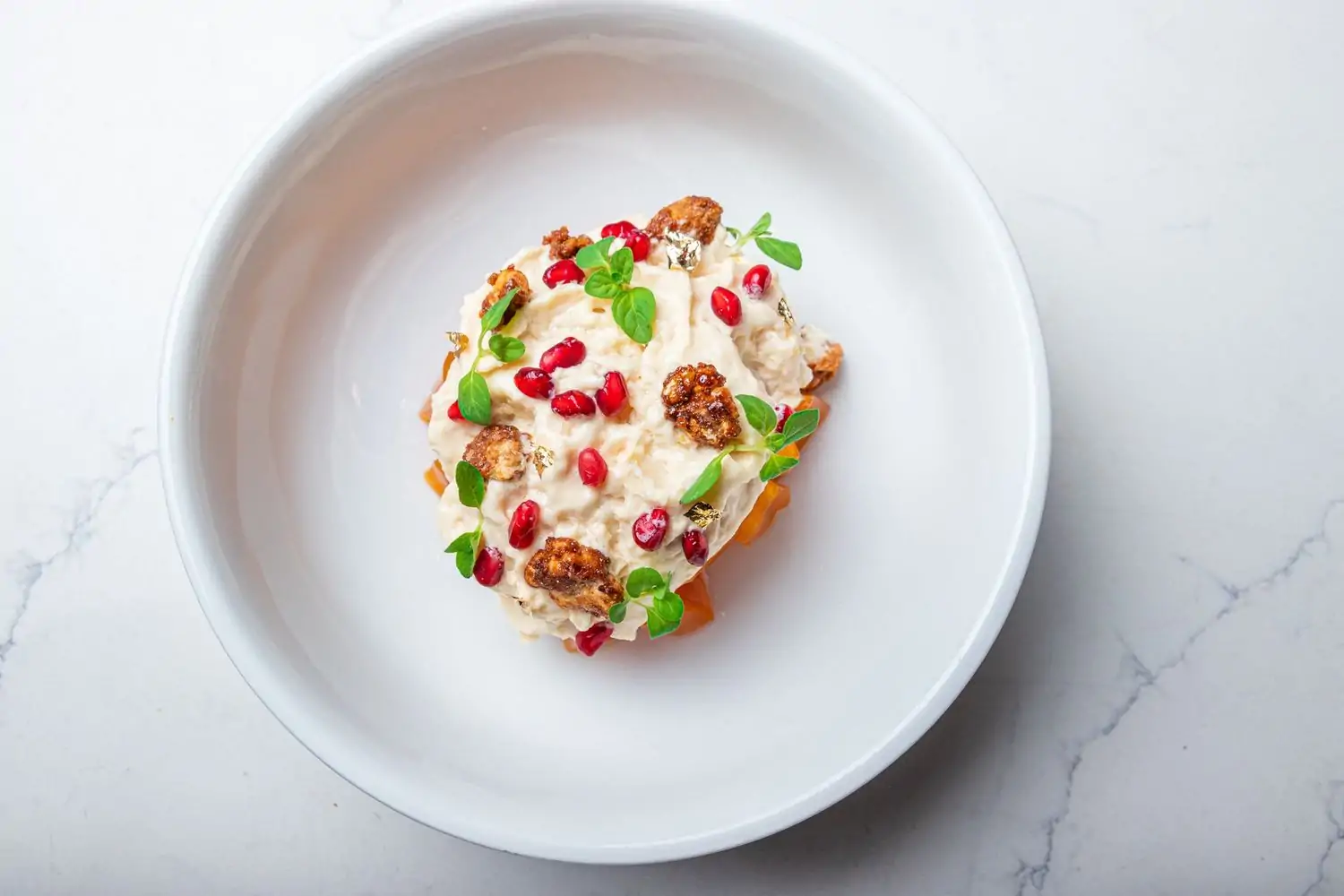 A plated dish from Doya, one of the best restaurants in Wynwood, Miami, with a creamy walnut sauce, pomegranate seeds, candied walnuts, fresh green herbs, and edible gold flakes on a crisp base, served in a white bowl on a marble surface—book on OpenTable.