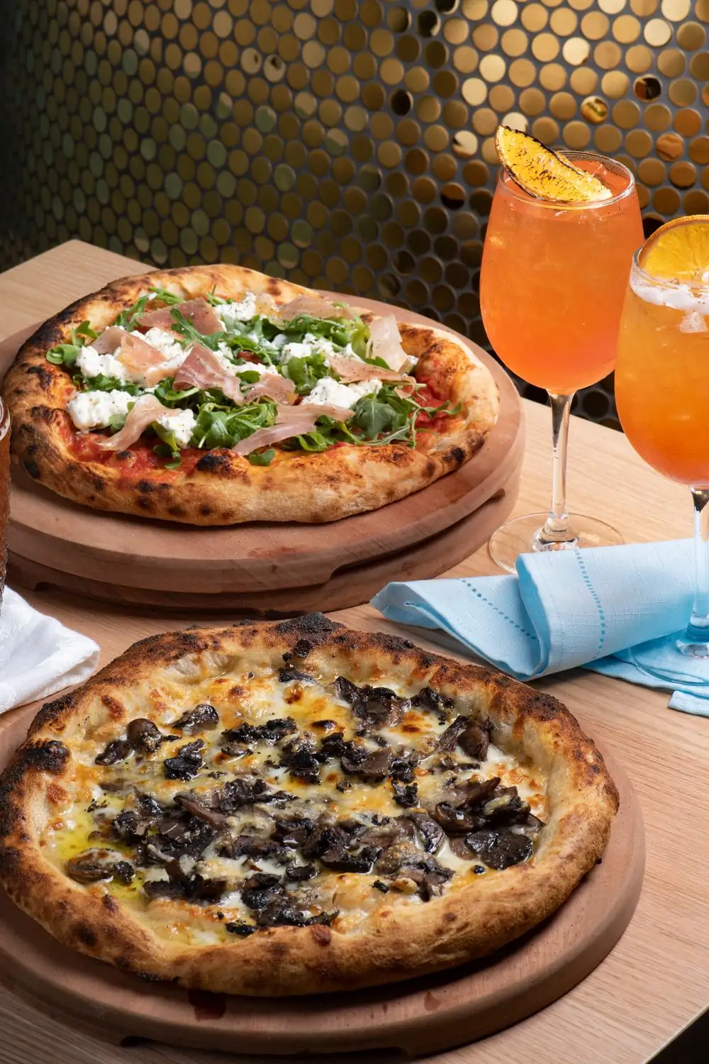 a view of two pizzas from Italica Midtown, one of the best restaurants in Wynwood, Miami. One pizza is topped with sliced ham, arugula, and ricotta cheese, while the other has sliced mushrooms. Also pictured are two Aperol spritz cocktails.