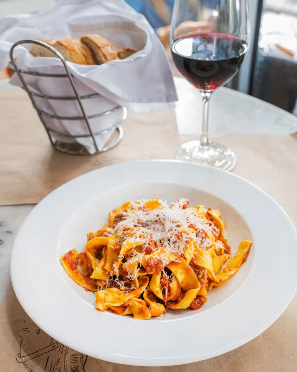 A plate of pappardelle pasta with meat ragù and grated cheese at Salumeria 104, one of the best restaurants in Wynwood Miami, accompanied by a basket of bread and a glass of red wine on a marble table.