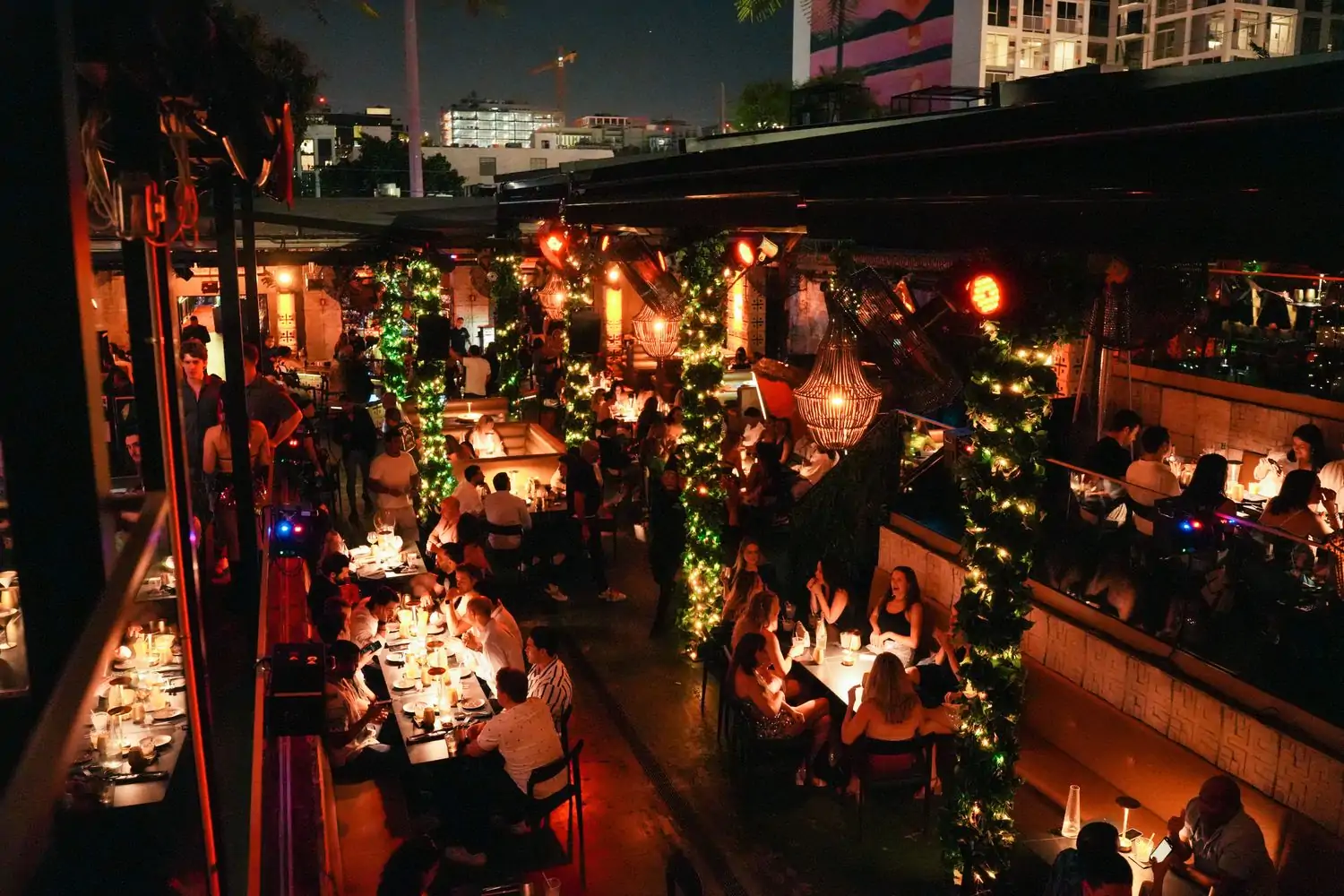 A lively nighttime dining scene at Mayami, one of Wynwood Miami's best restaurants, with warmly lit tables, lush greenery-wrapped columns, hanging chandeliers, and a stylish crowd enjoying dinner and drinks in an open-air setting.