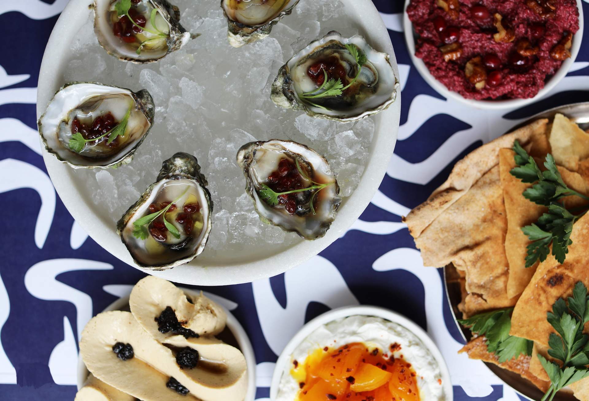 A plate of oysters on ice kept on blue tile next to a few dips including a red dip.