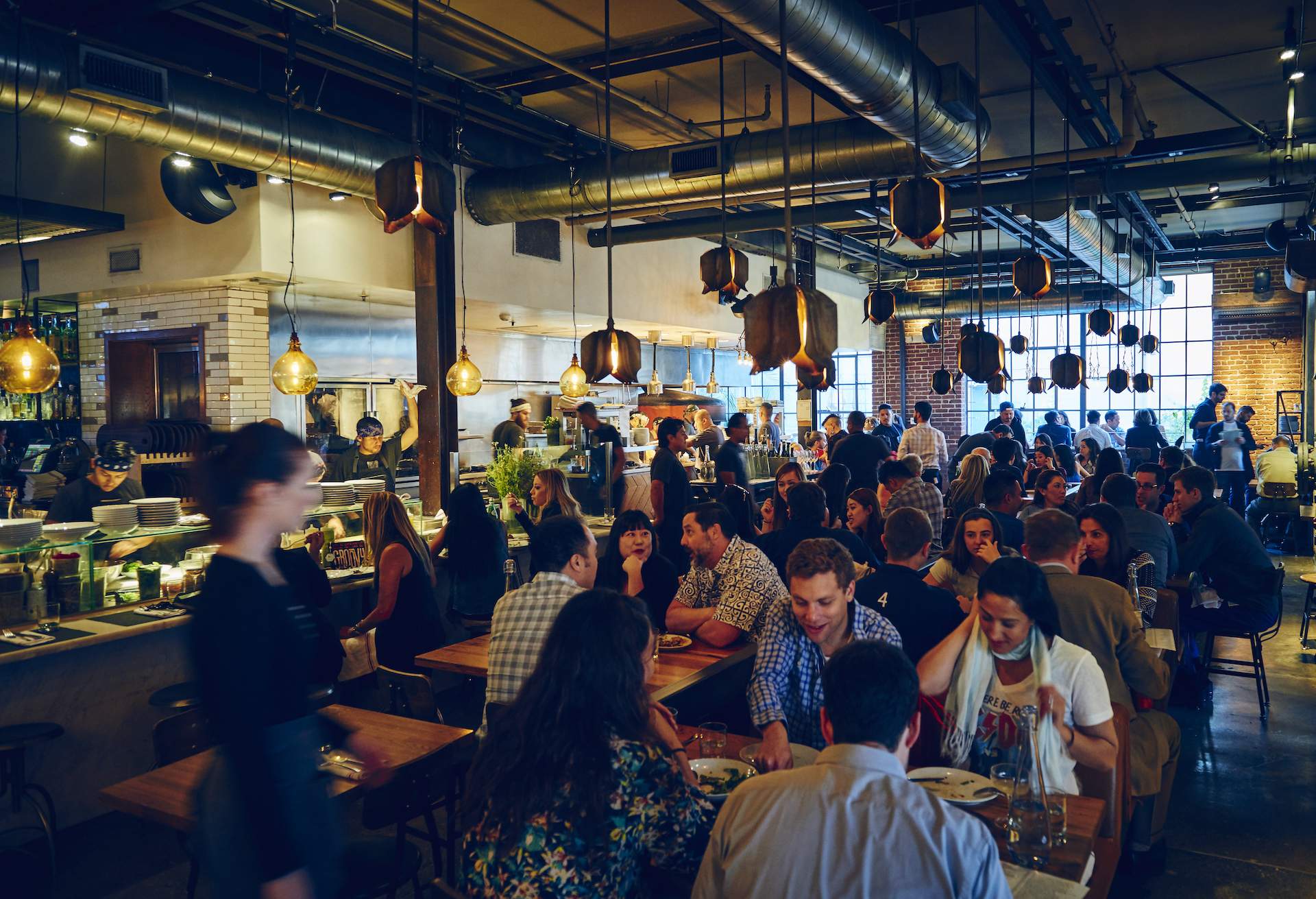 The interior of the LA restaurant Bestia which has low-hanging wooden ceilings, and lots of people sitting at wooden tables and chairs.