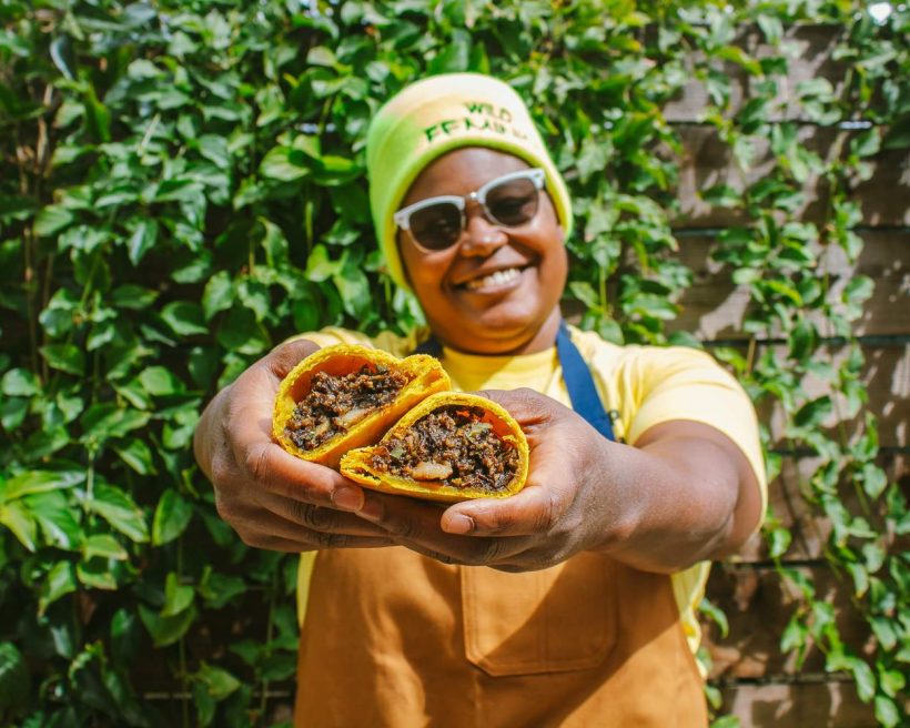 Rashida Holmes of Bridgetown Roti wears a green beanie while holding roti in front of her as she stands against a green background