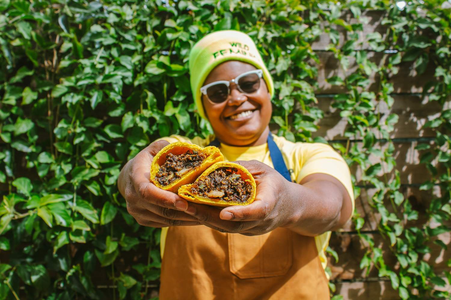 Rashida Holmes of Bridgetown Roti wears a green beanie while holding roti in front of her as she stands against a green background