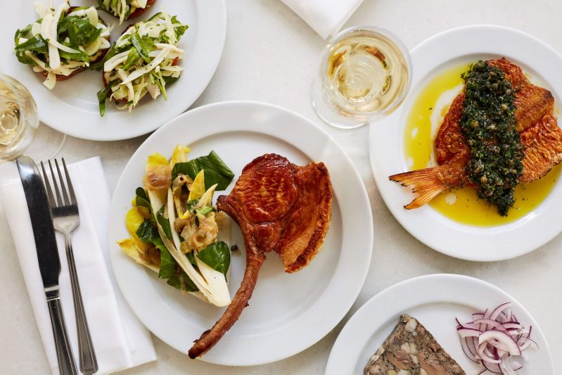 Plates with vegetable toast, a large cut of meat, and a whole fish on a white table cloth.
