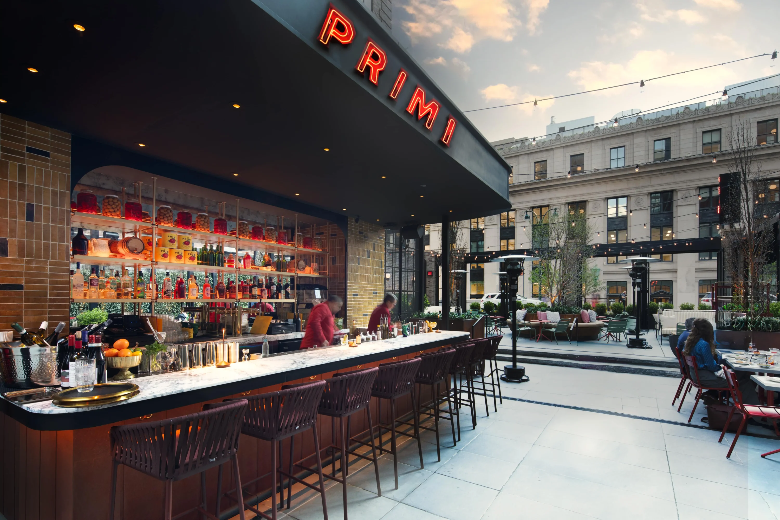 A backlit bar and burgundy bar stools across Moynihan Train Hall at Bar Primi in New York City