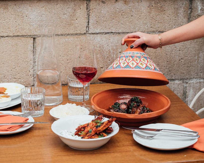 A hand lifts the top of a tagine decorated with a geometric pink and blue pattern containing braised wagyu beef cheek tagine at Bavel in Los Angeles