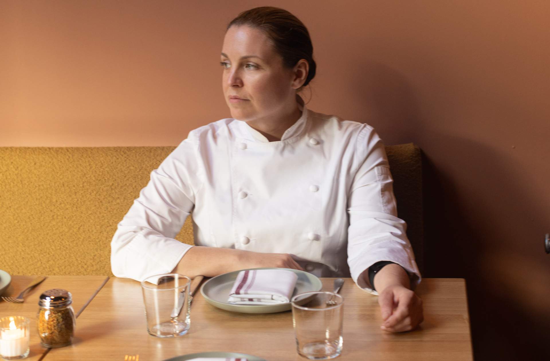 Chef Melissa Rodriguez in a white chef's coat sitting on a yellow bench set against a pink wall at her NYC restaurant Al Coro