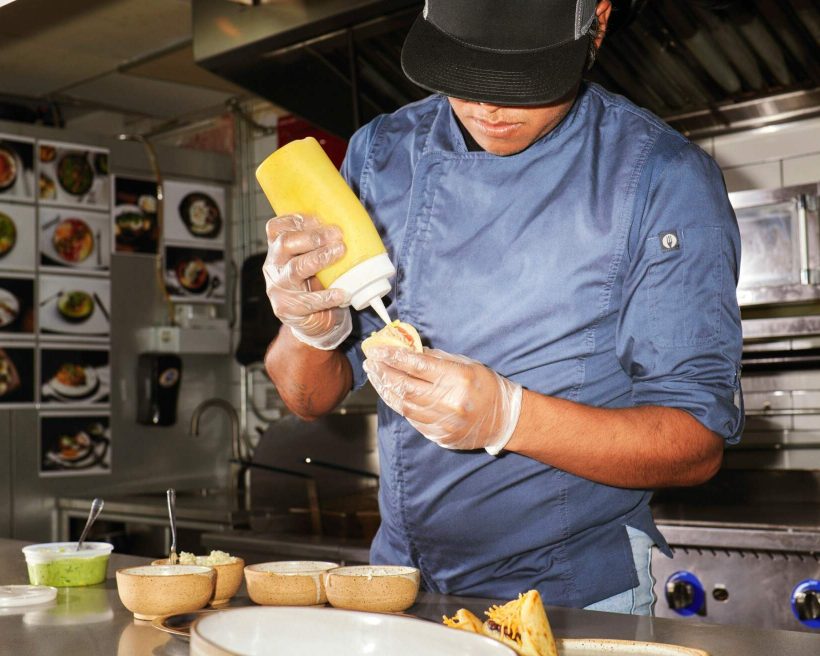 A chef in a blue chef's coat with a black hat pouring a squeezy bottle with yellow liquid in it on a sandwich. This image is being used to represent a story about mental health in the restaurant industry.