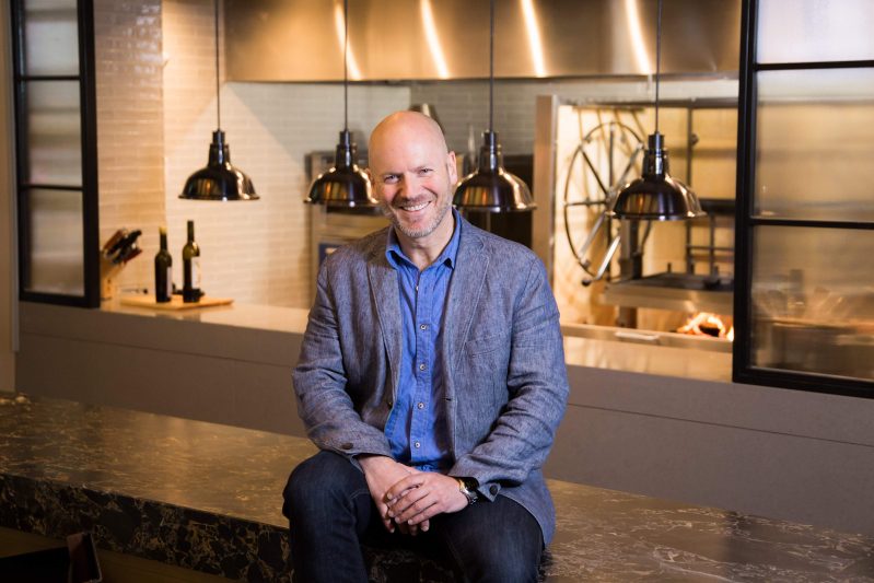 Chicago restaurateur Emmanuel Nony in a dark blue shirt and a light blue jacket sitting on a marble table at one of his restaurants.