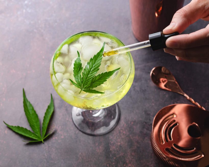 A hand holds a dropper of marijuana oil over a cannabis cocktail garnished with a cannabis leaf