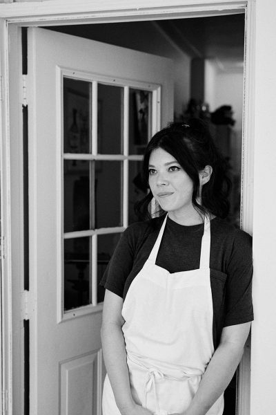 Natasha leans against a doorway while wearing a chefs apron