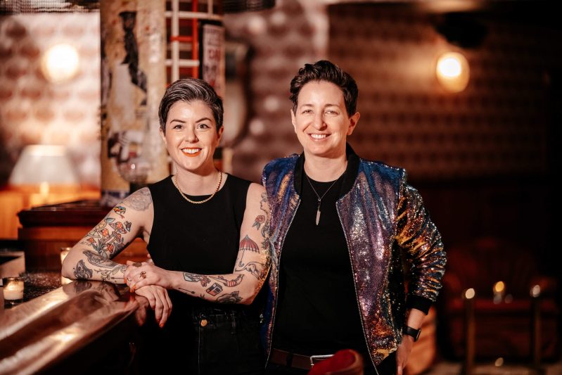 Chicago chef Zoe Schor in a sparkly purple jacket standing next to her fiancée Whitney LaMora in a black tank top.