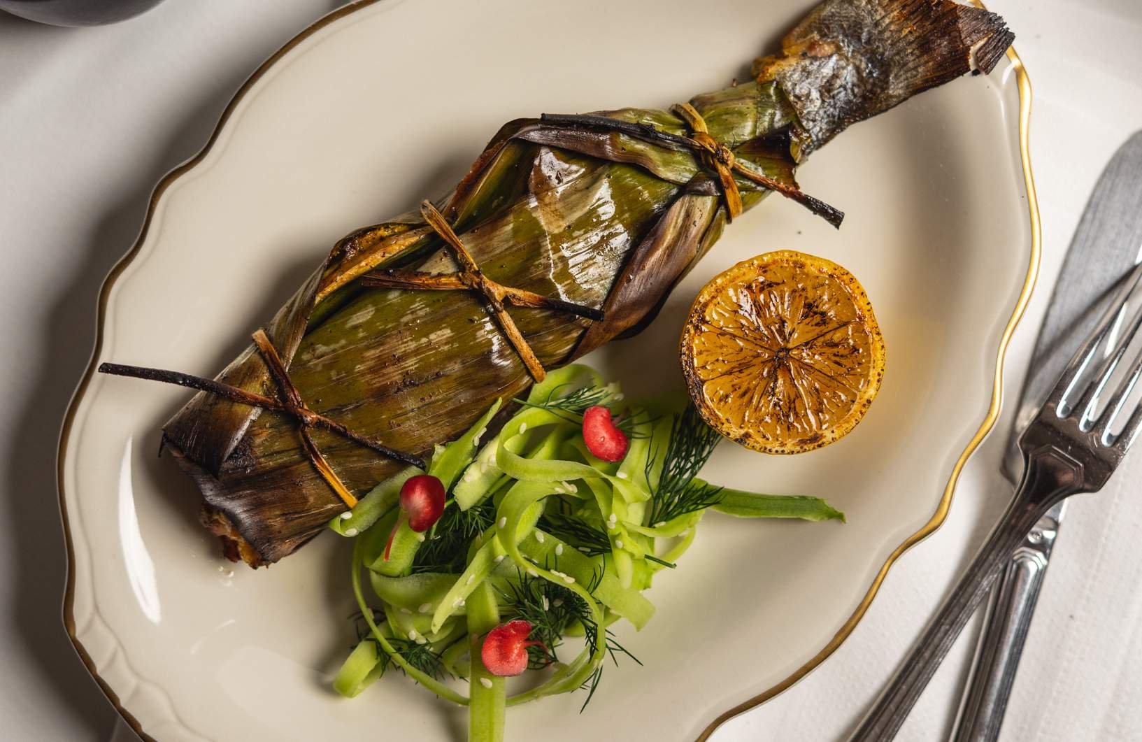 A piece of fish wrapped in a banana leaf that’s sitting on an oval-shaped white plate along with a grilled, sliced lemon and a small side salad at NYC Indian restaurant Sona.