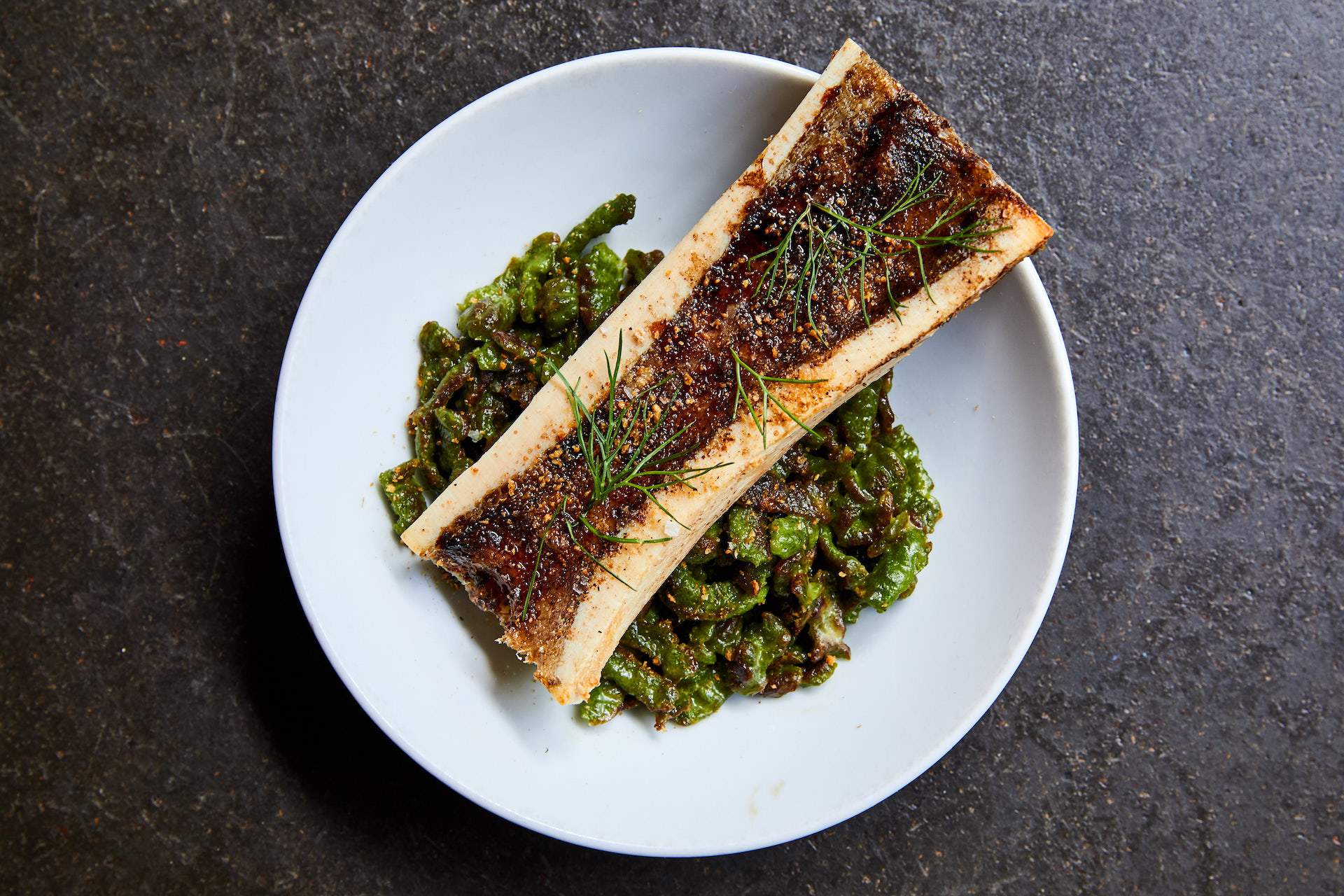 Roasted bone marrow balancing on the rim of a white plate above spinach gnochetti, smoked breadcrumbs, and aged balsamic at Bestia restaurant in Los Angeles.