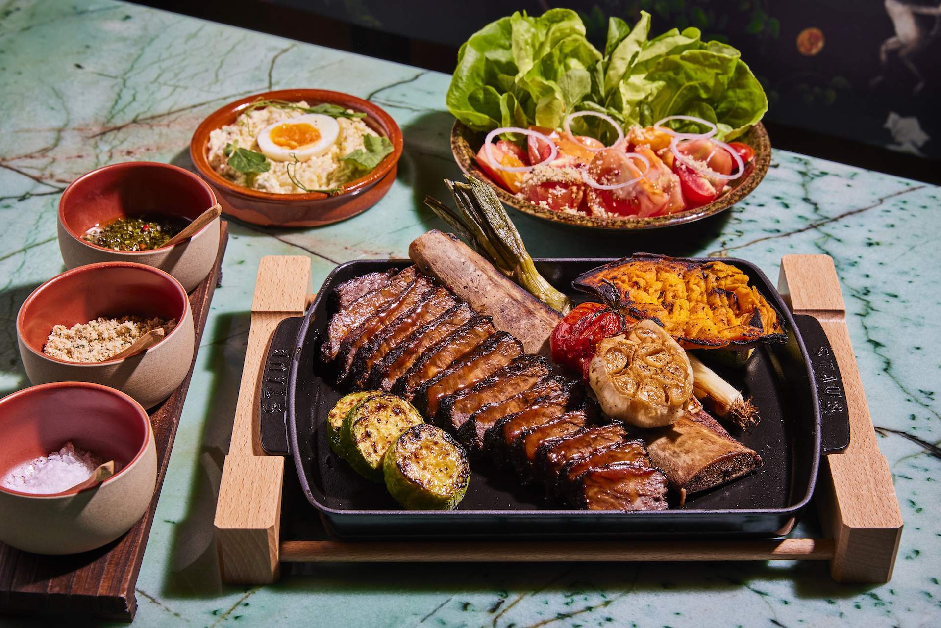 Sliced meats in a black vessel surrounded by small plates of food on a marble table at Los Angeles restaurant Qué Bárbaro