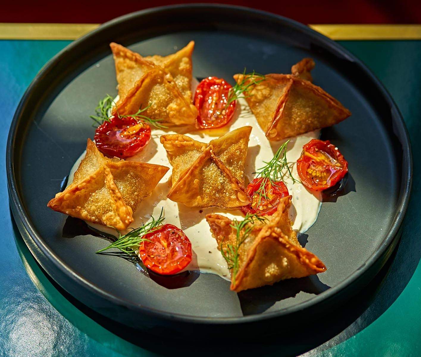 Fried dumplings called manti on a black plate with sliced tomatoes and a white sauce underneath at New York City restaurant Zou Zou’s.