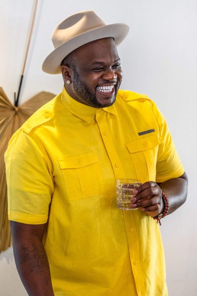 A man wearing a yellow shirt and a wide brimmed hat holds a cocktail and smiles.