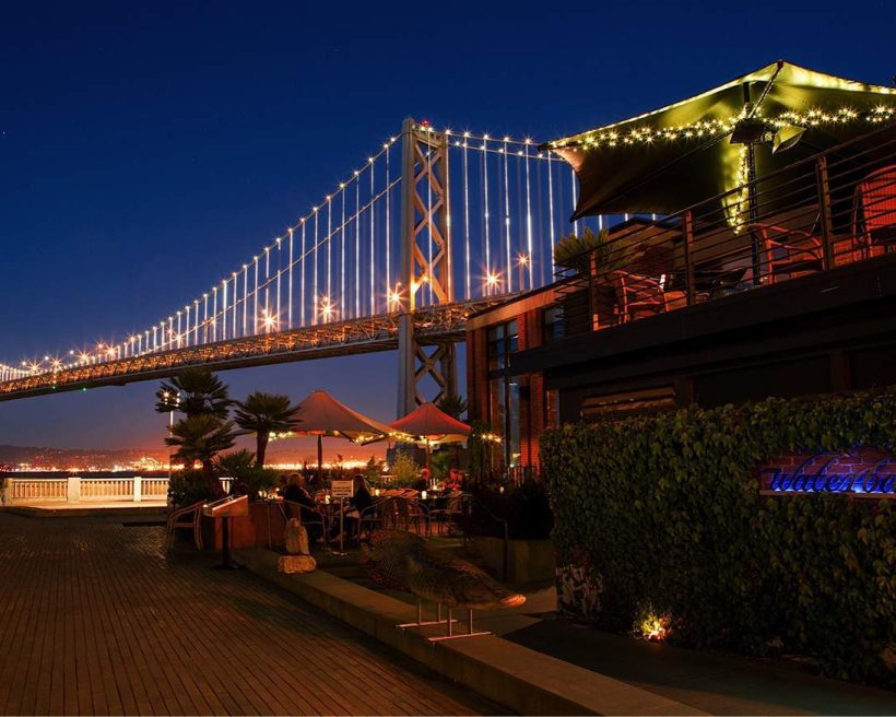 The waterfront outdoor seating of San Francisco restaurant Waterbar with the Bay Bridge lit up in the background.