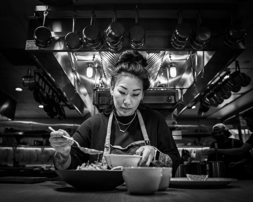 A female chef wears an apron and spoons sauce over a bowl.