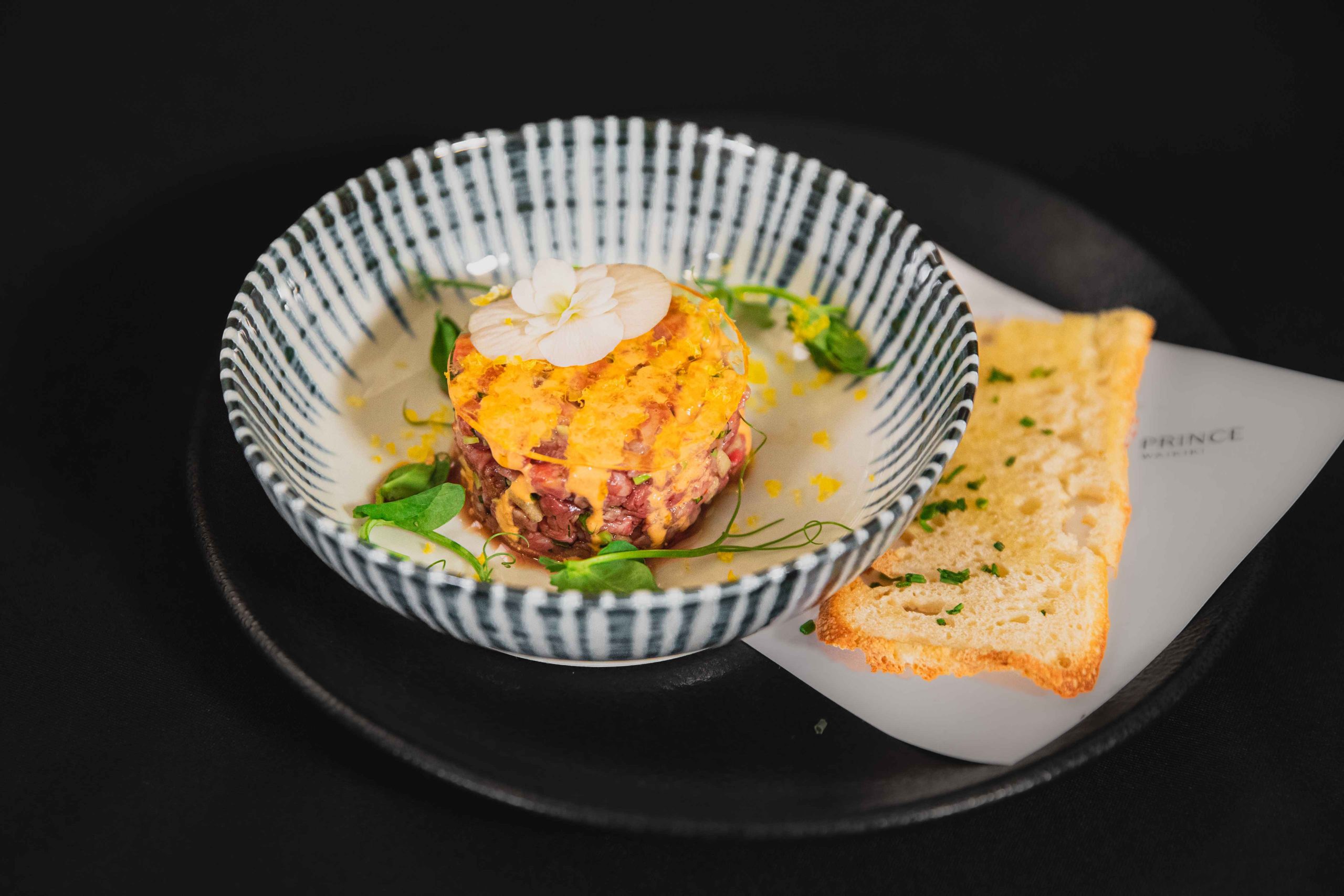 A black and white striped ceramic bowl with American wagyu, mayonnaise, hot sauce, tamari, edamame, apple, and cured egg yolk at 100 Sails Restaurant & Bar in Honolulu
