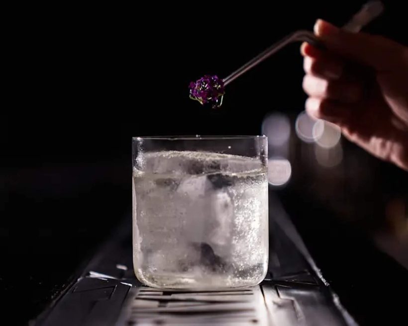 A hand shown holding a pair of tweezers placing a flower in a clear cocktail at Mexico City cocktail bar Handshake Speakeasy