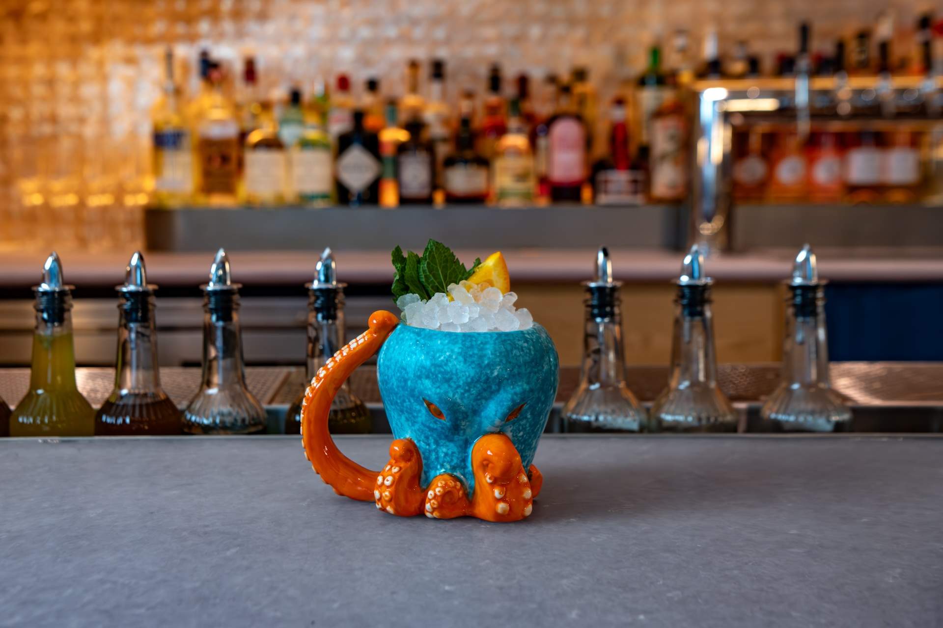 A cocktail in a tiki-style mug shaped like an octopus with a blue body and orange tentacles