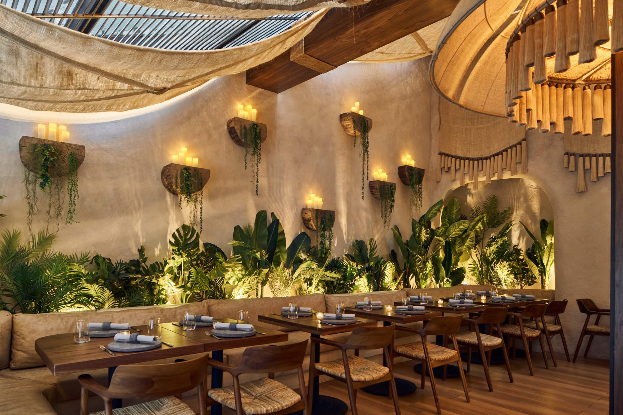 The interior of NYC Mexican seafood restaurant Tán featuring sconces with candles on the walls, tropical plants, and wooden tables