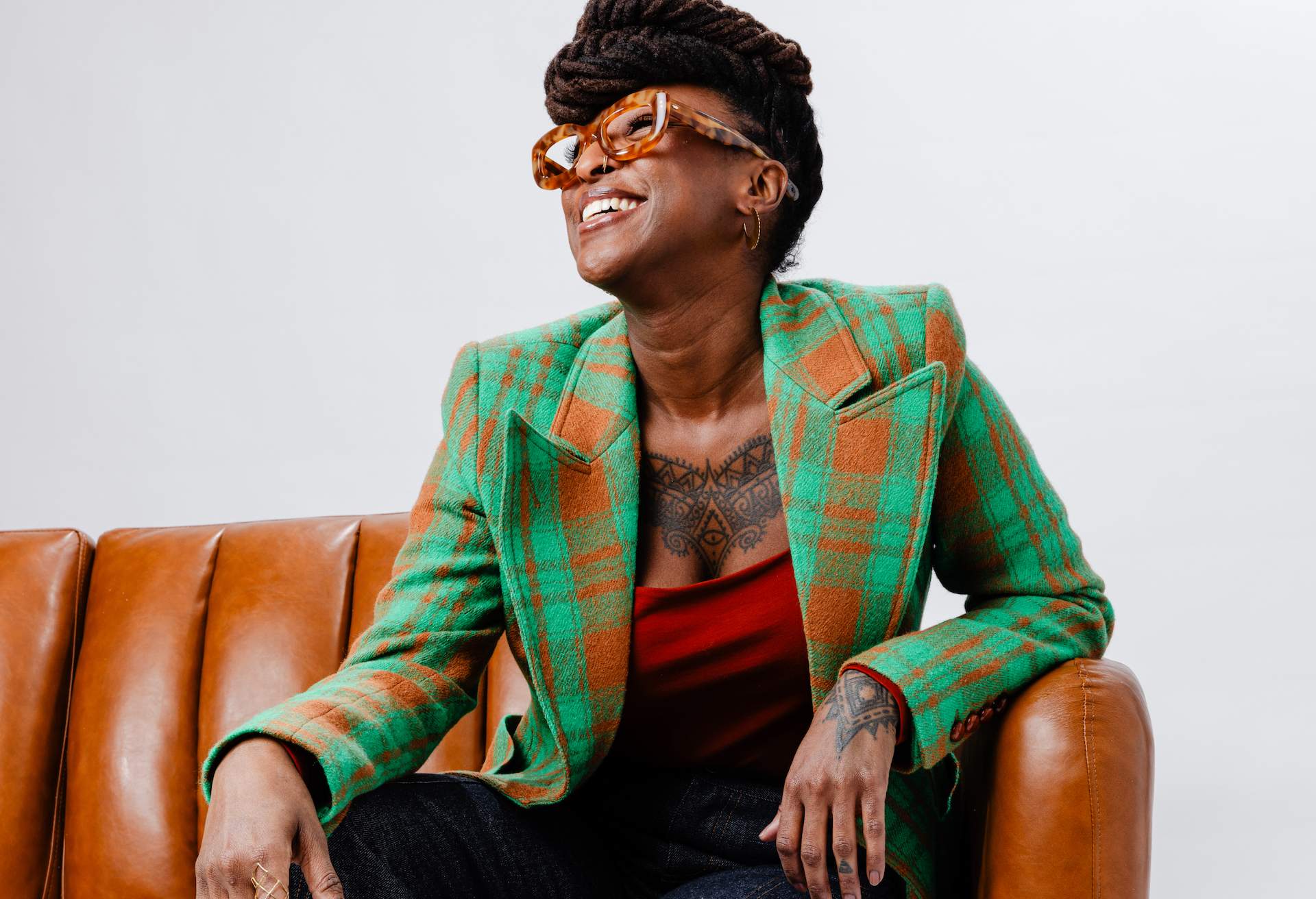 Chef Mariya Russell sits on a leather couch wearing a green and orange blazer, looking off to the side and smiling.