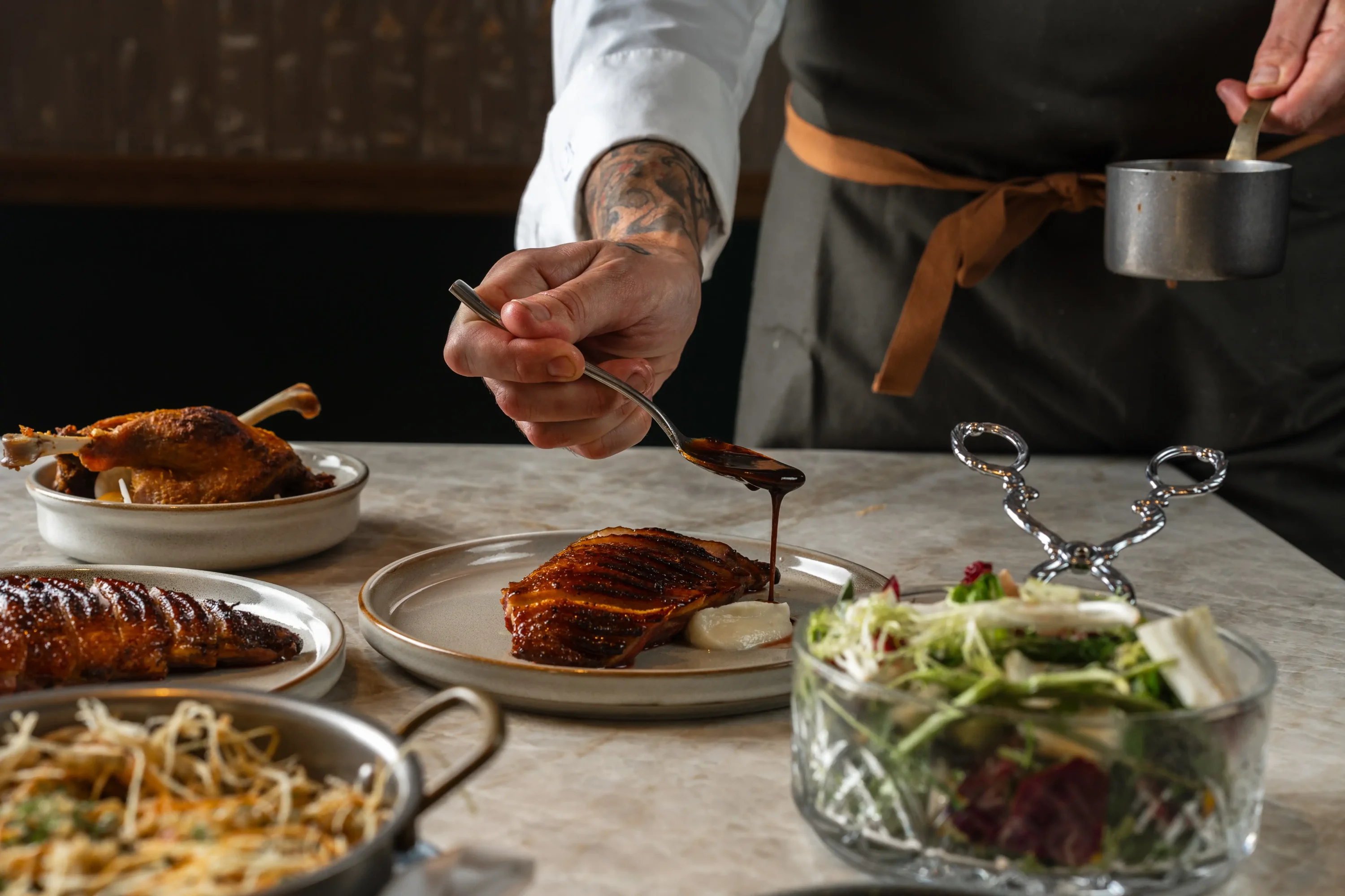 A man in a black apron pouring sauce on meat surrounded by a salad and other dishes.