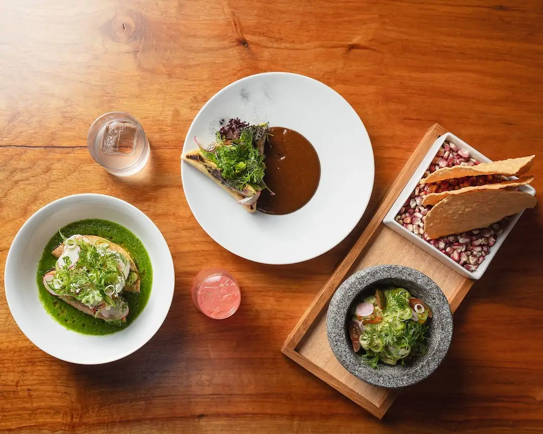 A table with two cocktails, shrimp aguachile in tortillas, mole sauce, and greens at Mexta in Austin. Book on OpenTable.