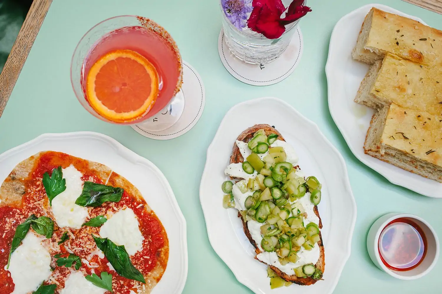 A table setting with focaccia, pizza, and bread surrounded by vibrant cocktails at The Tailor’s Son, one of the best Italian restaurants in San Francisco.