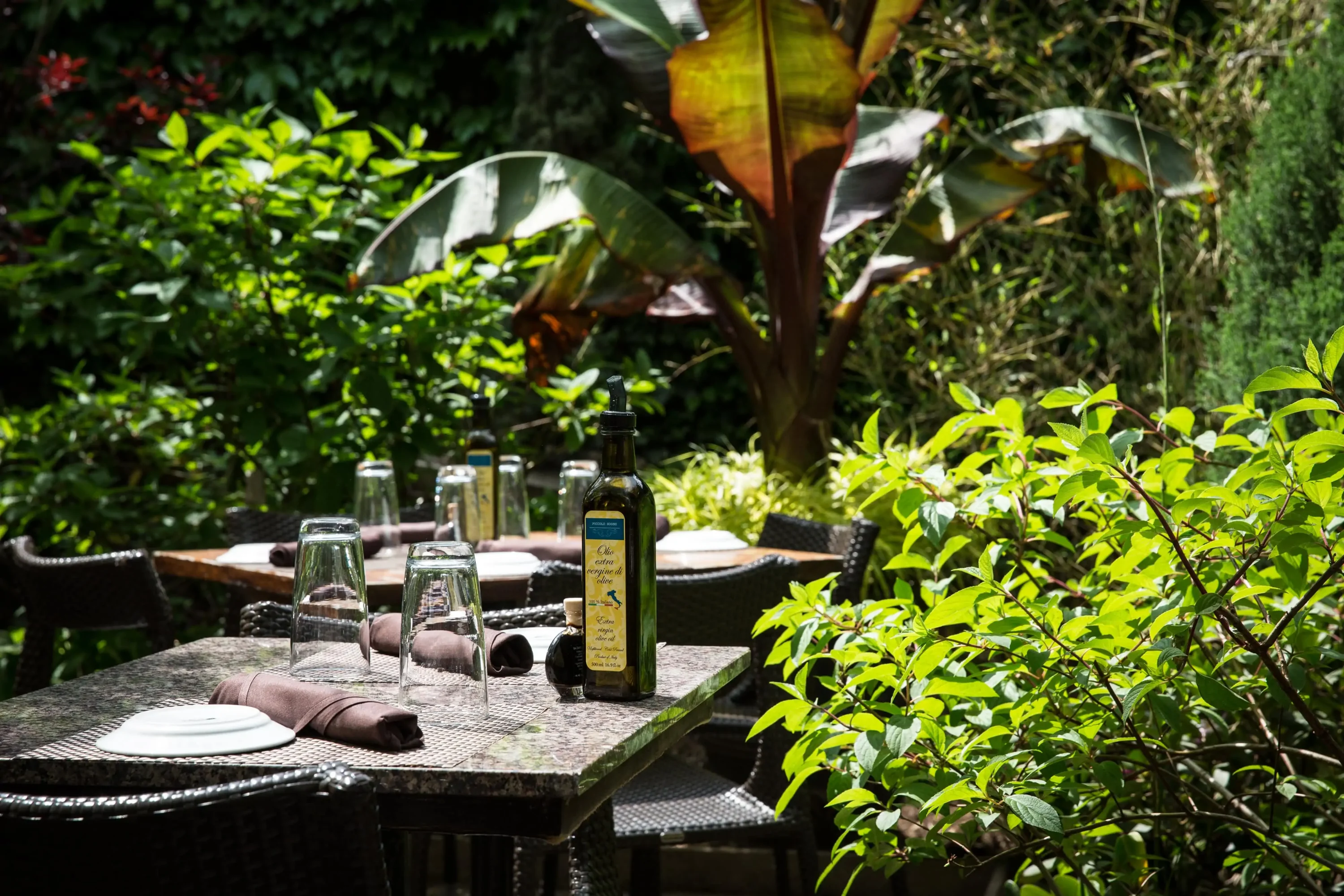 The corner of an outdoor patio features two tables with table settings and an olive oil bottle surrounded by lush plants and trees at Piccolo Sogno in Chicago. Book on OpenTable.