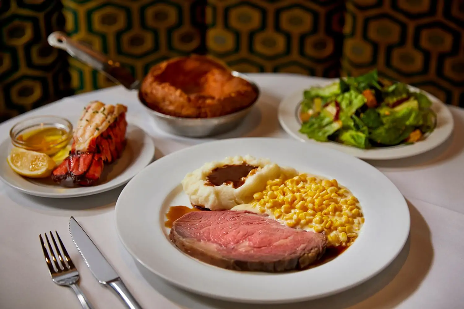 A table setting featuring a plate of prime rib with corn and mashed potatoes and gravy, with a side salad and a pan on the table next to the main plate at Lawry’s The Prime Rib - Beverly Hills, one of the best restaurants in Beverly Hills, Los Angeles. Book on OpenTable.