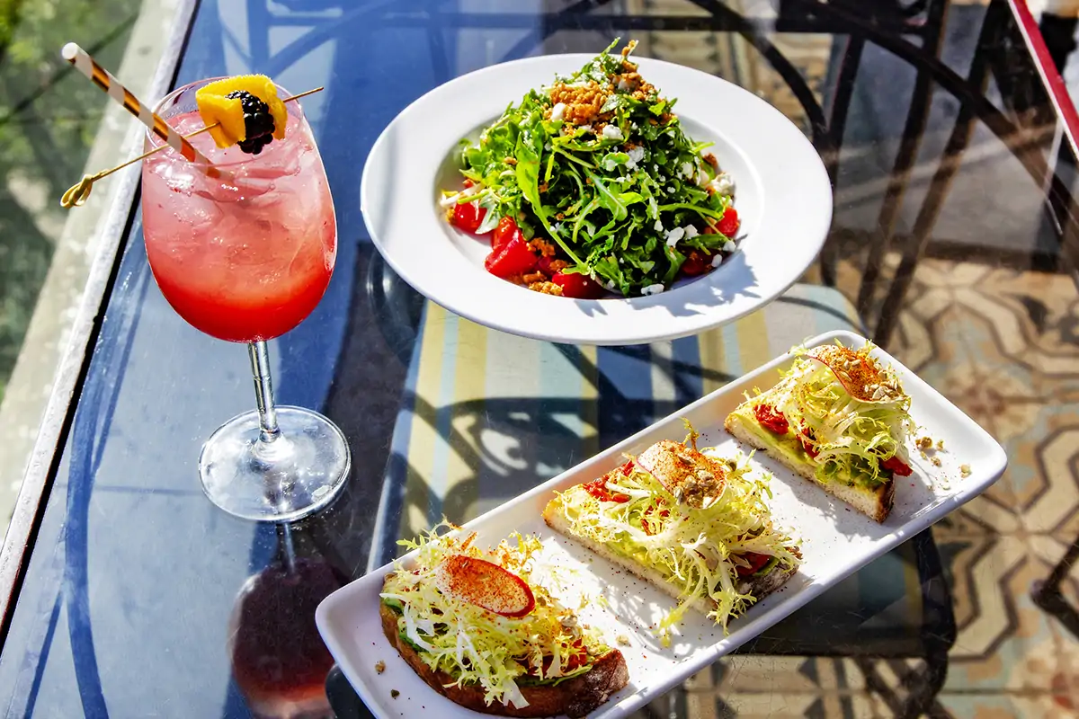 A spritz with a white and gold straw, a white plate with a salad with tomatoes, and a rectangular white plate with three pieces of toast topped with avocado and frisée at Perch LA, one of the best restaurants in Downtown LA. Book on OpenTable.