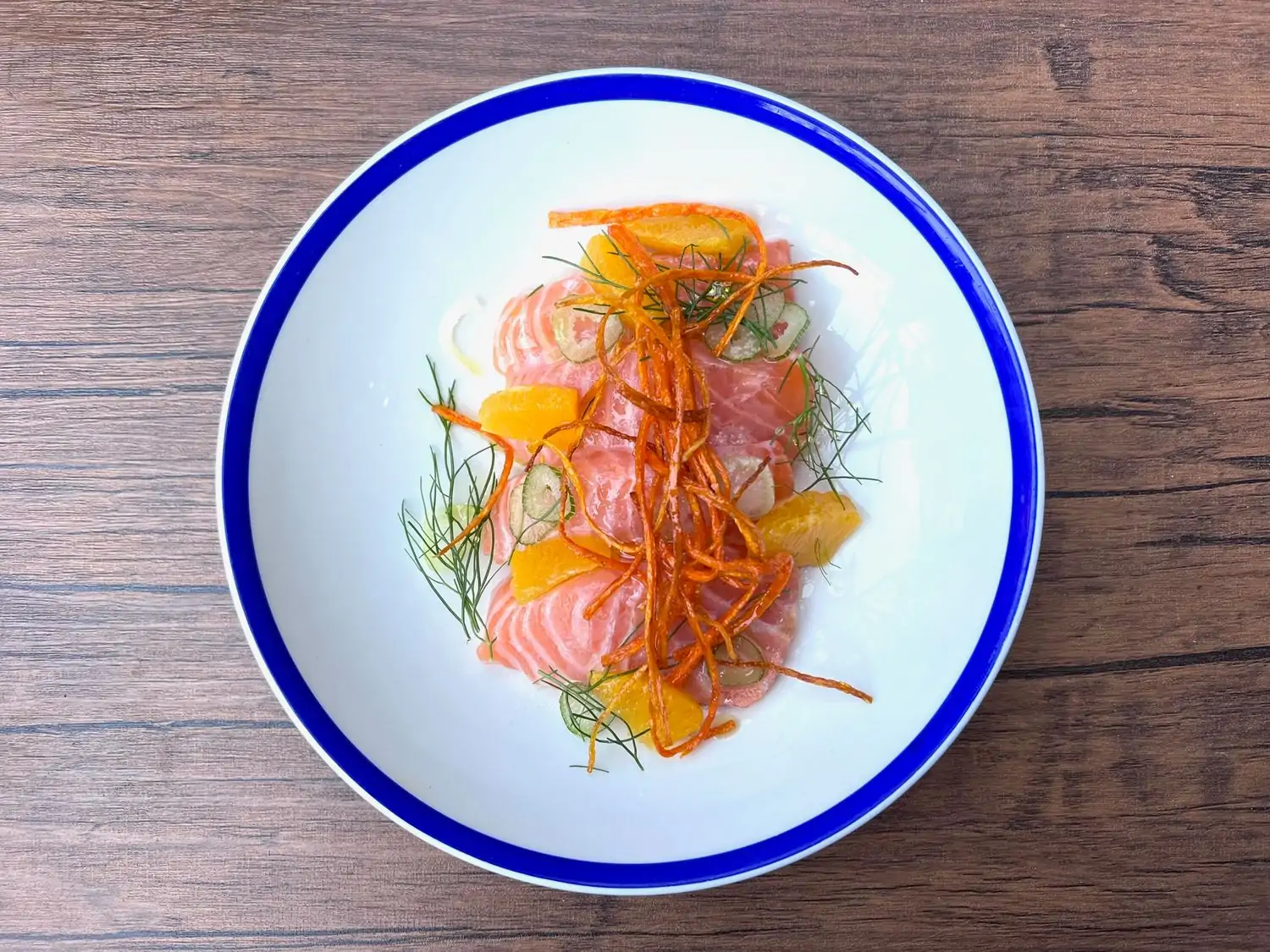 A white plate with a blue rim with raw fish topped with herbs and orange at Little Whale in Boston. Book on OpenTable.