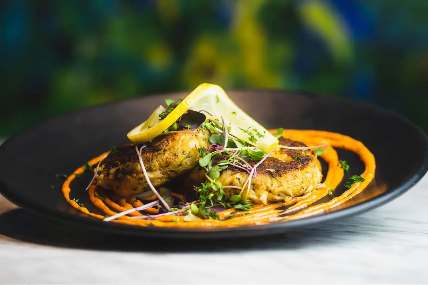 Crab cakes cooked to a golden brown are surrounded by stripes of orange sauce and topped with greenery and lemon at Kitchen + Kocktails, one of downtown Dallas's best restaurants, available on OpenTable.